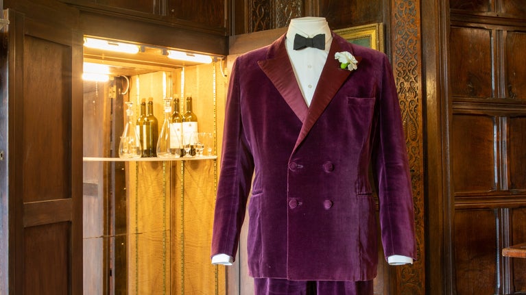 Top half of Lord Fairhaven's wine-coloured velvet smoking suit on the right and white carnation in the pocket, with a cupboard open behind it and to the left, with a light on and wine bottles on a shelf. Brown panels in room.